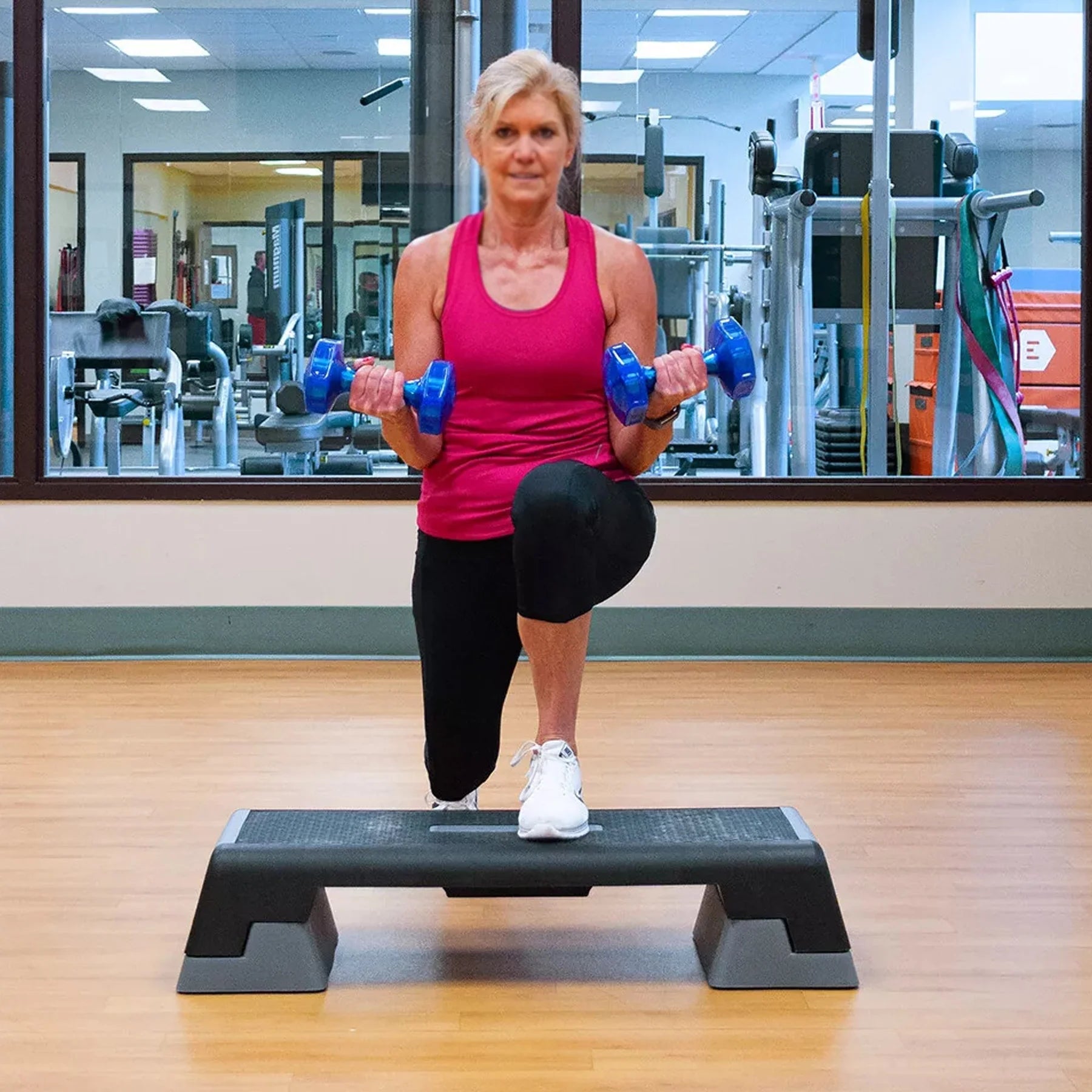 Woman exercising with blue dumbbells on a step platform in a gym.
